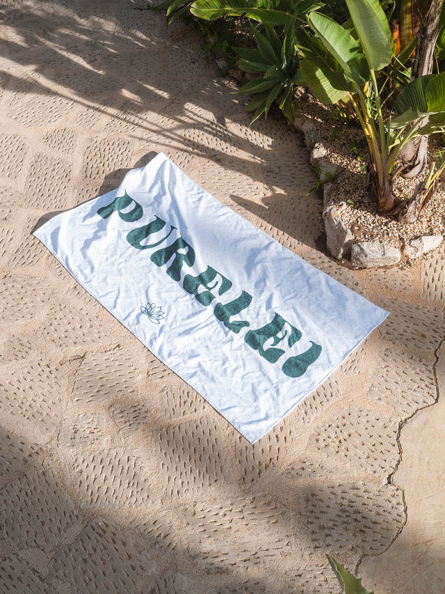 Weißes PURELEI Beach Towel Shoreline aus Baumwolle mit grünem Schriftzug, am Strand in natürlichem Licht liegend.