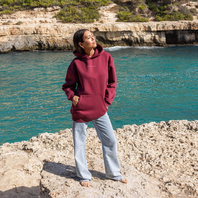 Locker sitzender weinroter PURELEI Endless Summer Hoodie aus Baumwollmix am felsigen Meer in entspannter Stimmung