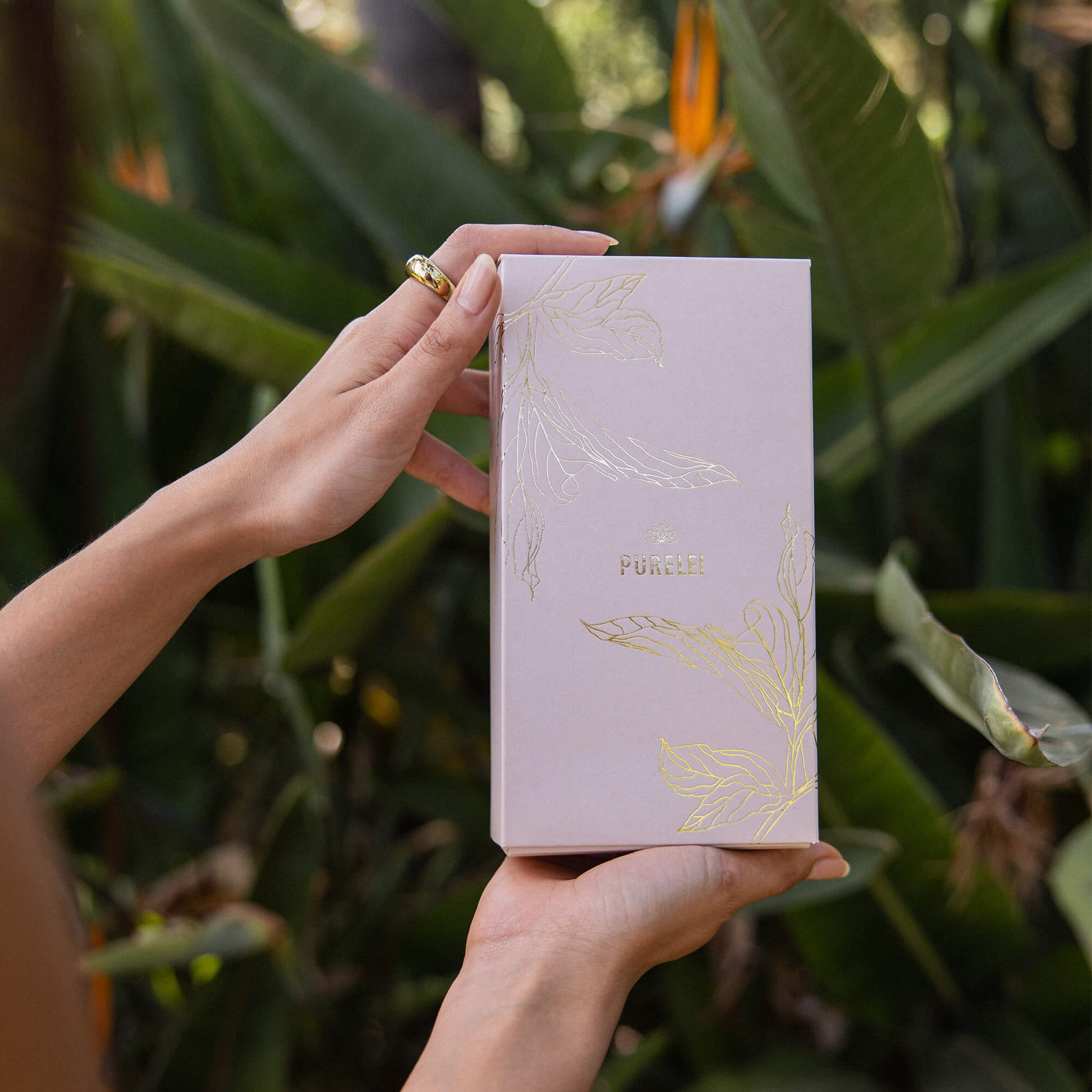 Elegante PURELEI Geschenkbox in zartem Rosa mit goldenen Akzenten, umgeben von tropischer Natur.