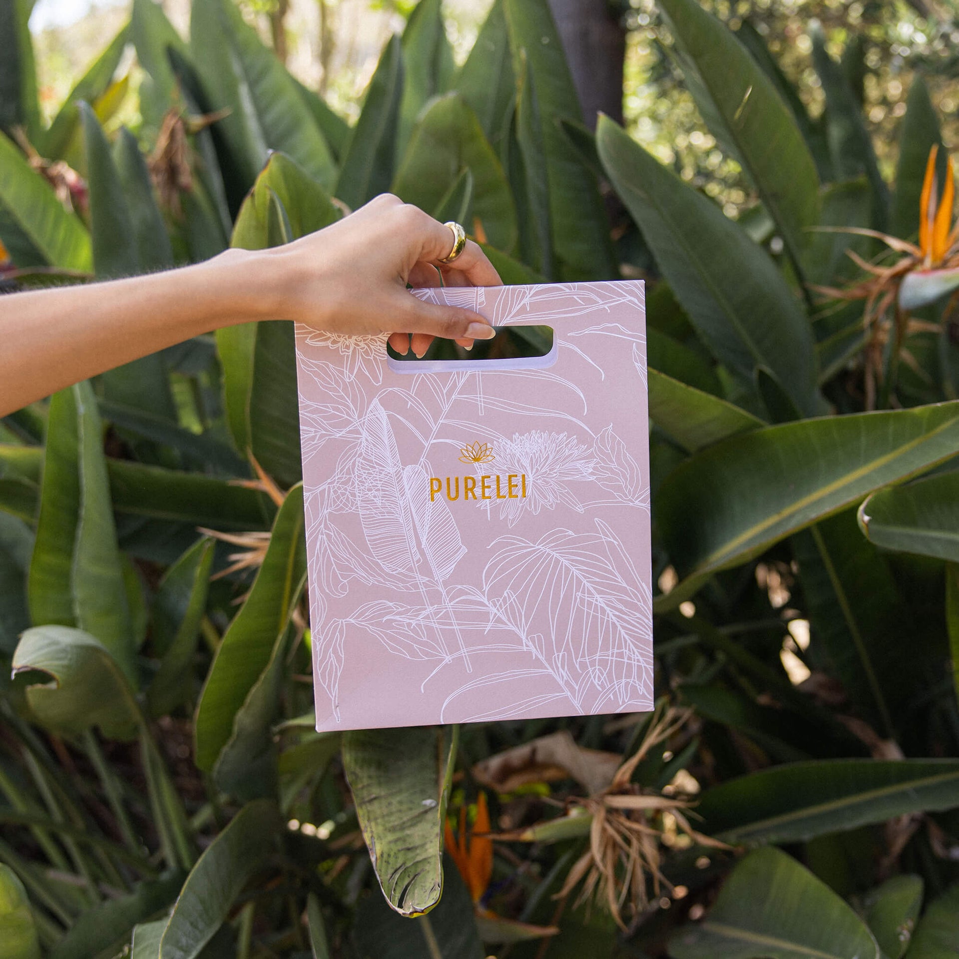 Lavendelfarbene PURELEI-Tüte mit goldener Schrift, umgeben von tropischem Grün.