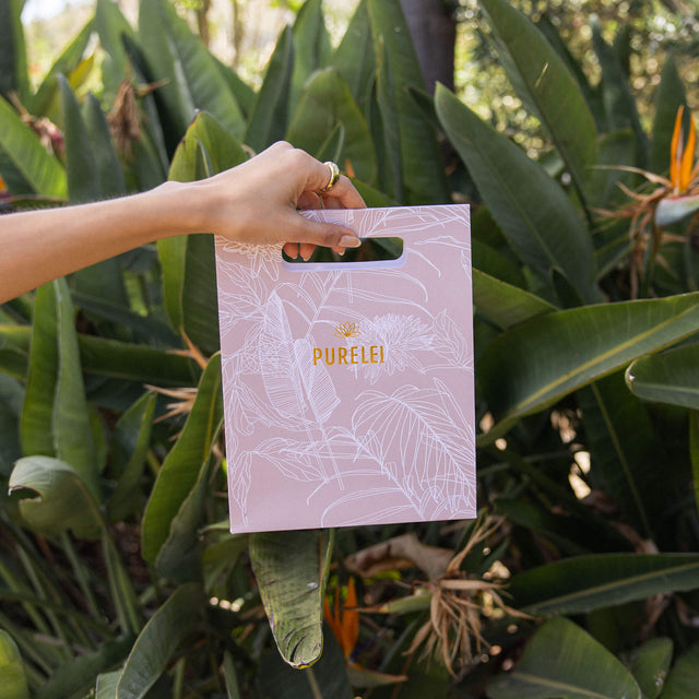 Lavendelfarbene PURELEI-Tüte mit goldener Schrift, umgeben von tropischem Grün.