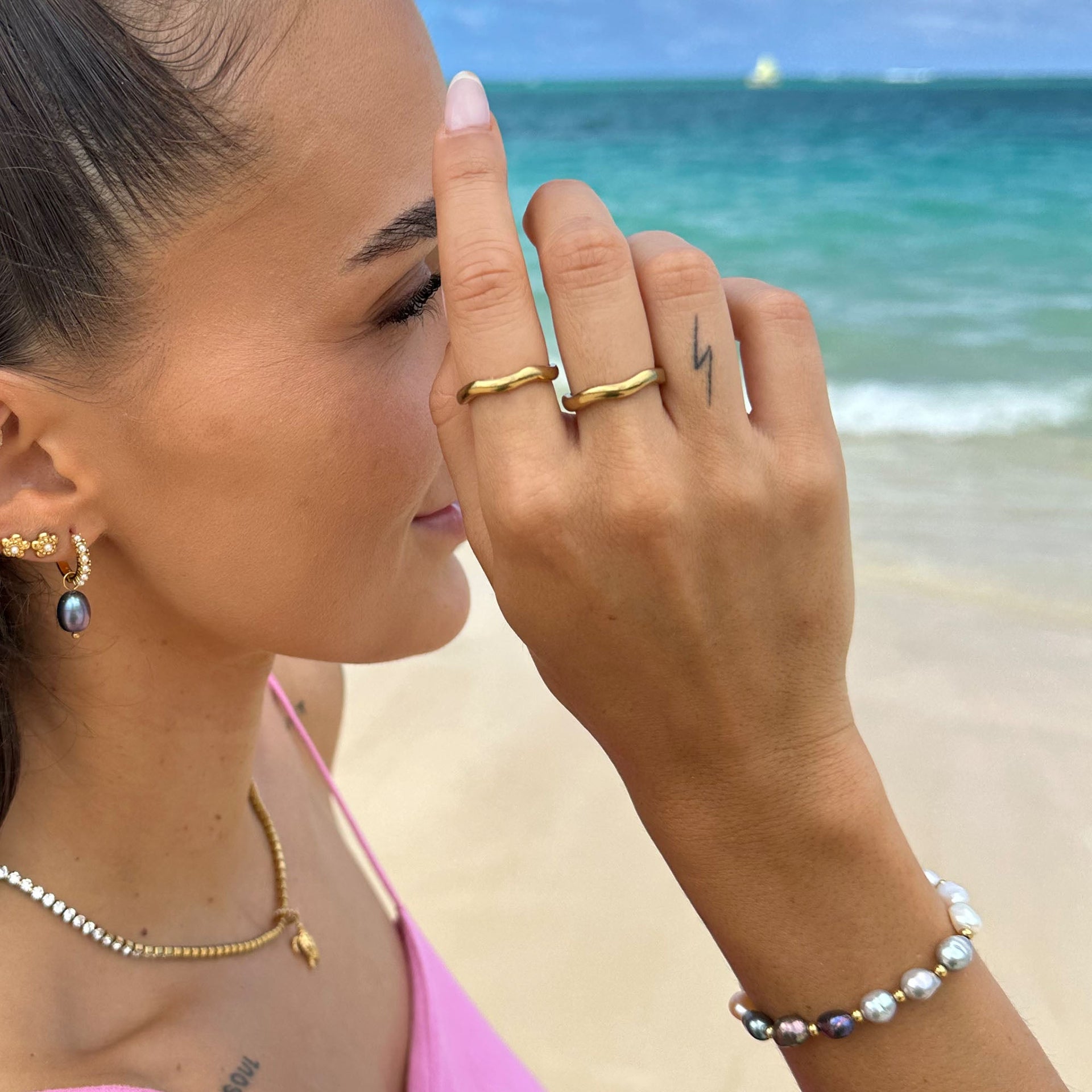 PURELEI Nakuna Ring in 18K Gold mit geschwungener Form, getragen am Strand vor türkisfarbenem Meer