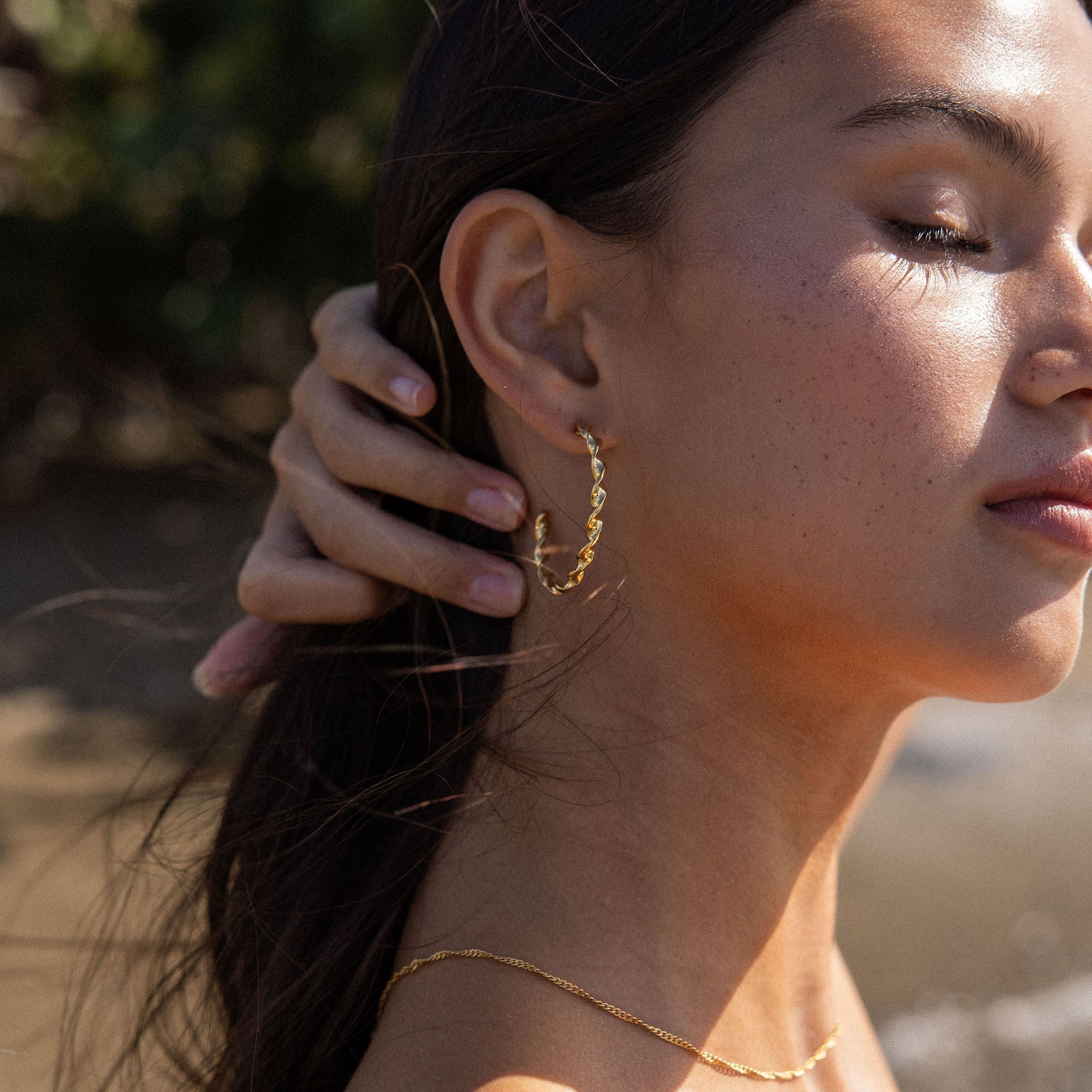 PURELEI Kaula Creole Ohrringe aus 18K Gold mit gedrehtem Design, sonnendurchflutet am Strand getragen.