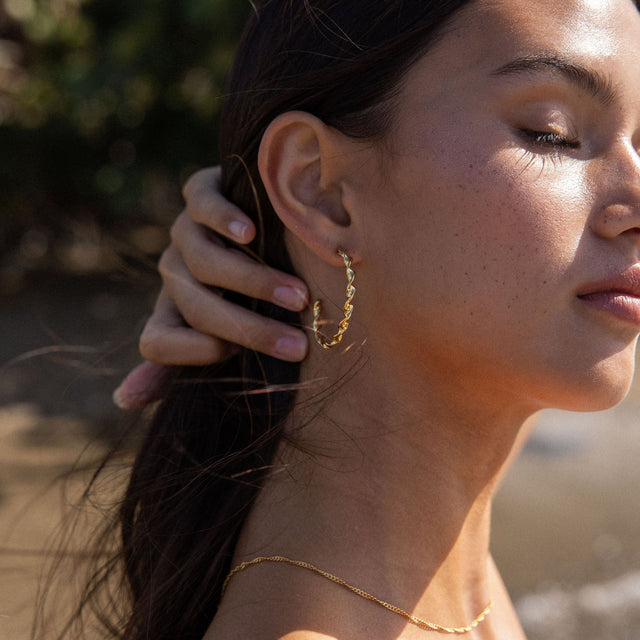 PURELEI Kaula Creole Ohrringe aus 18K Gold mit gedrehtem Design, sonnendurchflutet am Strand getragen.