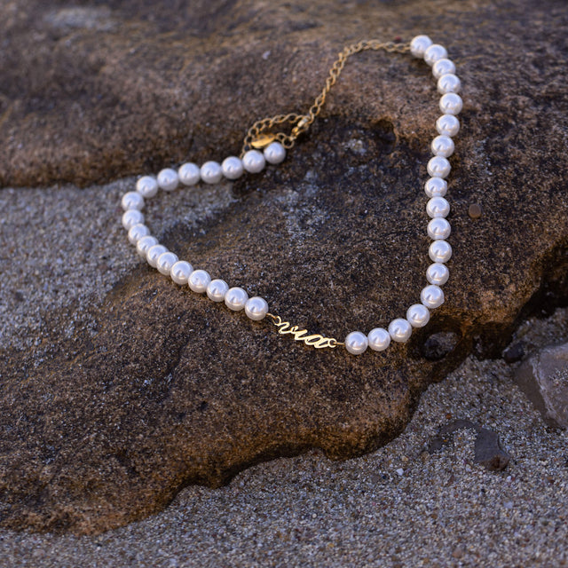 Via Pearl Perlenkette von PURELEI mit goldfarbenem via-Schriftzug und weißen Muschelperlen auf sandigem Stein.