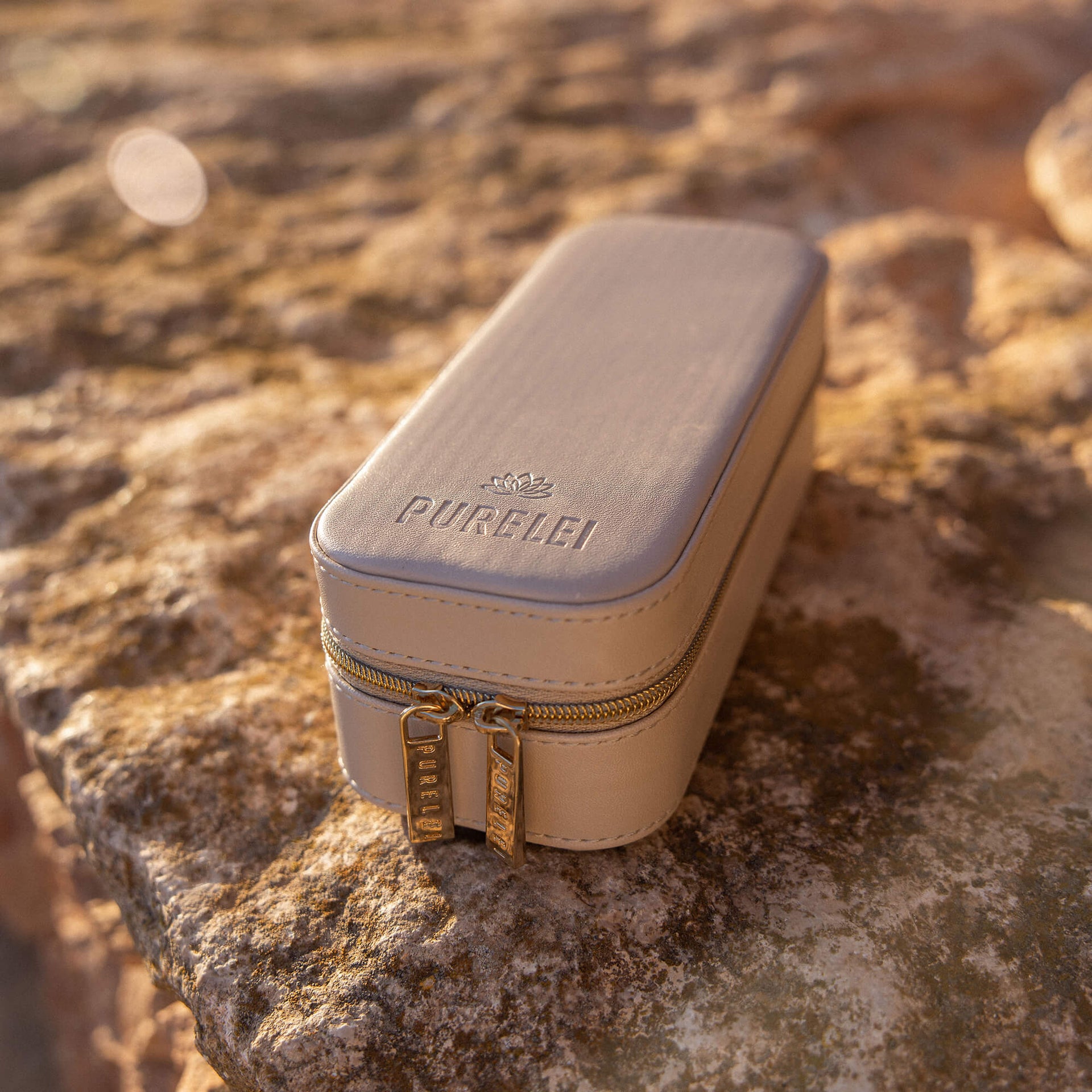 Beige Schmuckbox von PURELEI mit goldenen Reißverschlüssen auf steiniger Oberfläche.