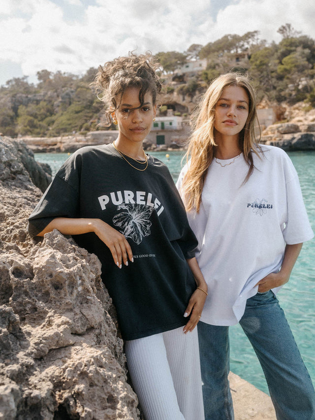 Zwei Frauen in PURELEI T-Shirts, eines schwarz mit Blumenmotiv, das andere weiß, am Wasser.