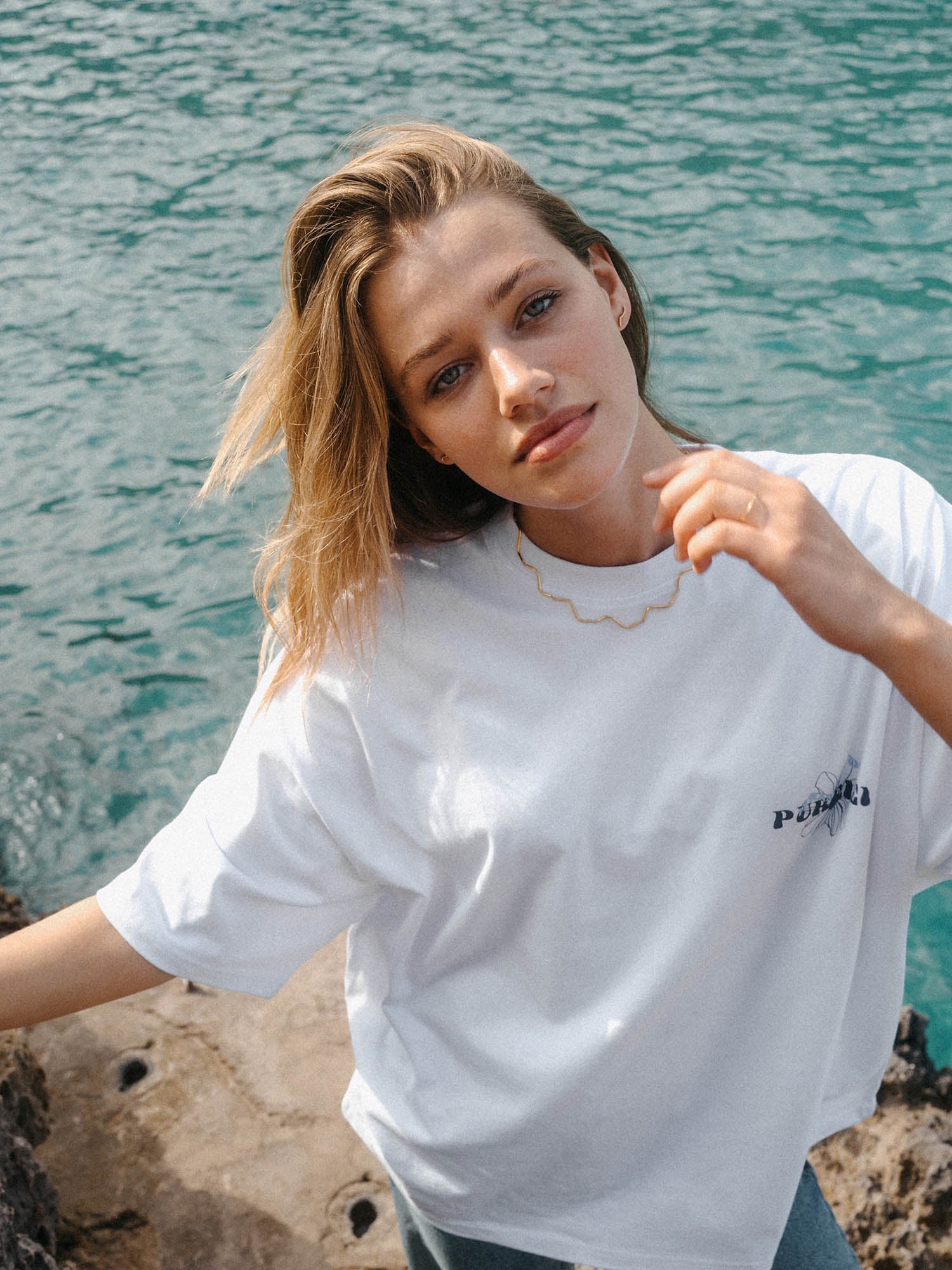 Roségoldene Wellenkette von PURELEI, getragen vor türkisfarbenem Wasser.