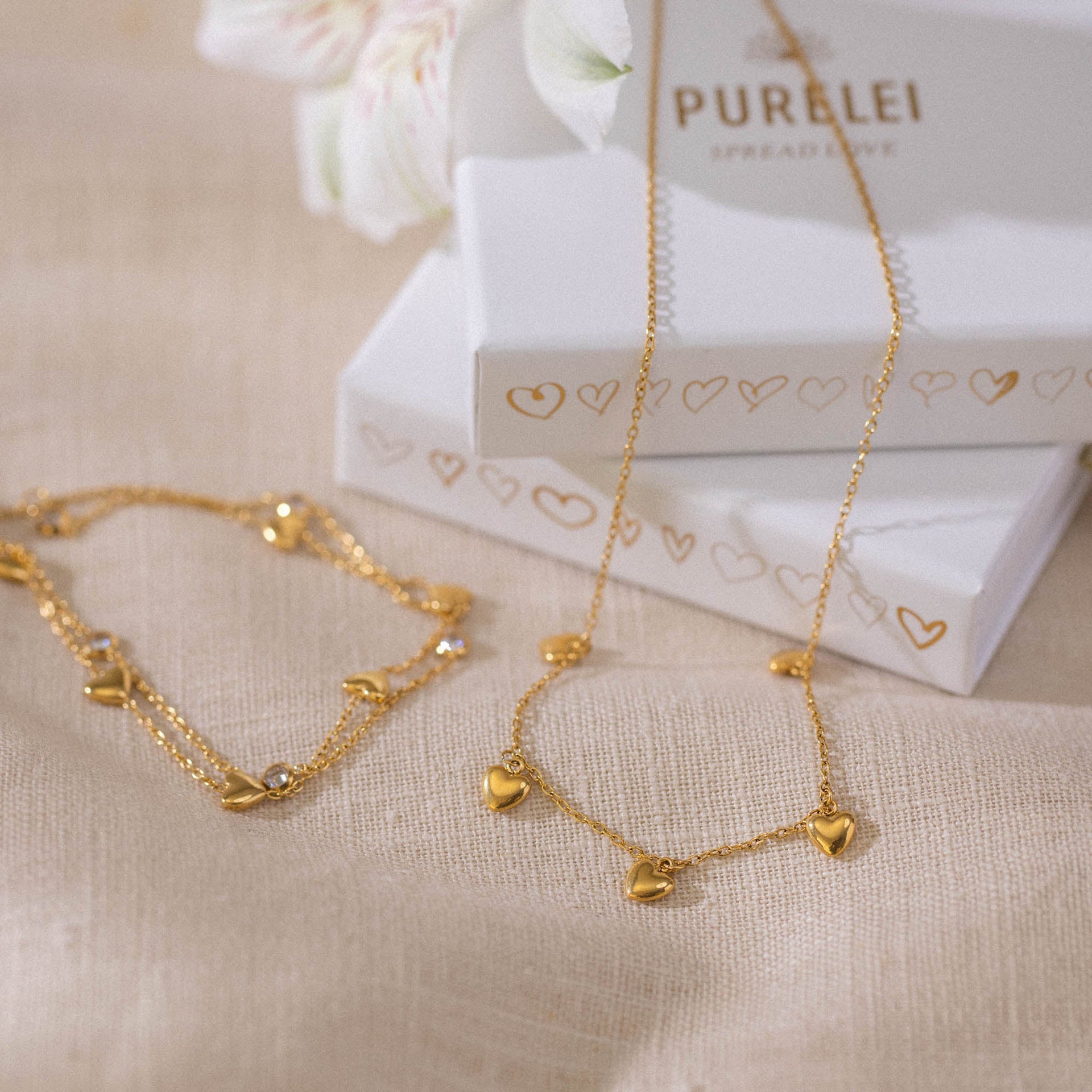 Goldene Kette und Armband mit Herzanhängern, elegant auf Leinen präsentiert – PURELEI.