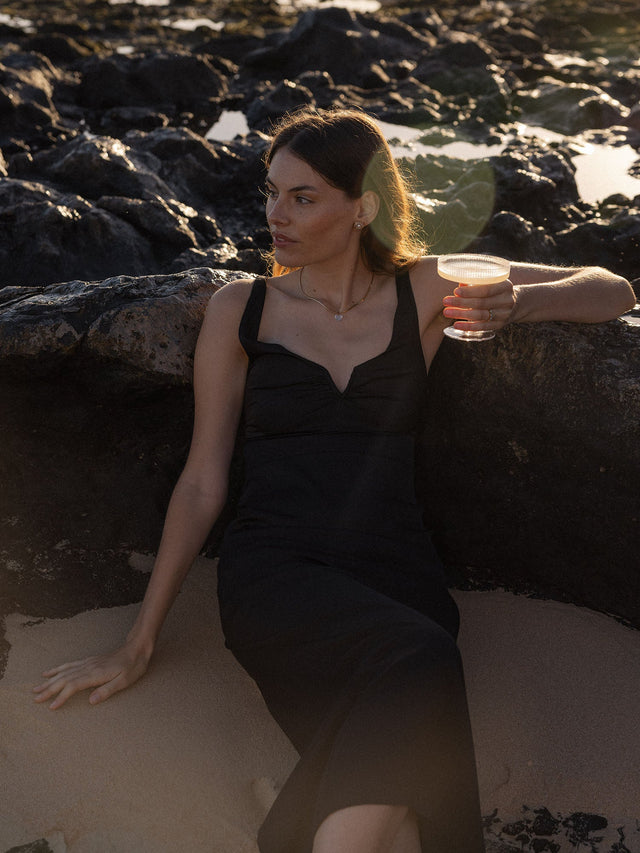 Elegantes schwarzes Kleid, getragen am Strand mit PURELEI Schmuck, entspannte Atmosphäre.