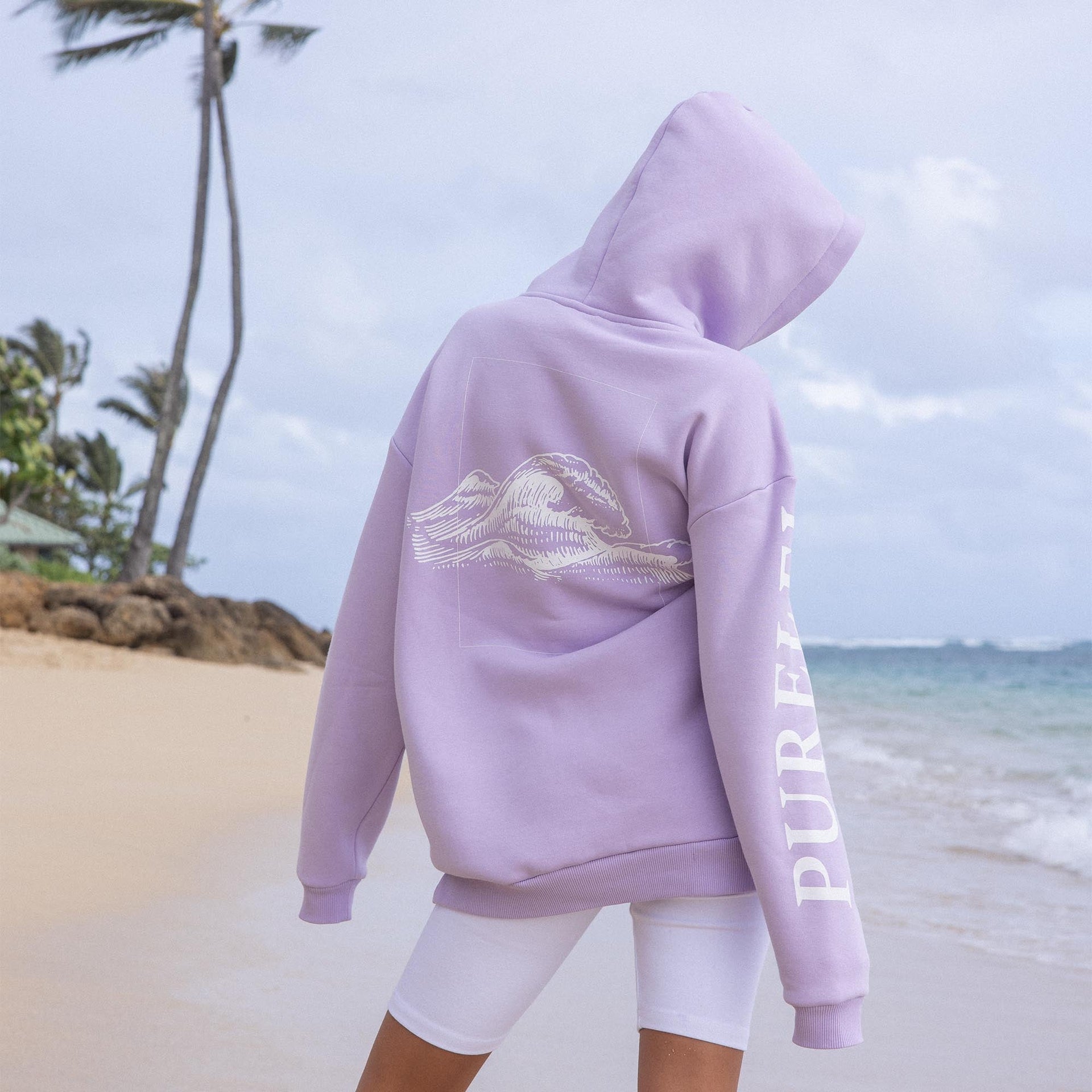 Lila Kapuzenpullover mit Wellenmotiv von PURELEI, am Strand getragen, entspannte Stimmung.
