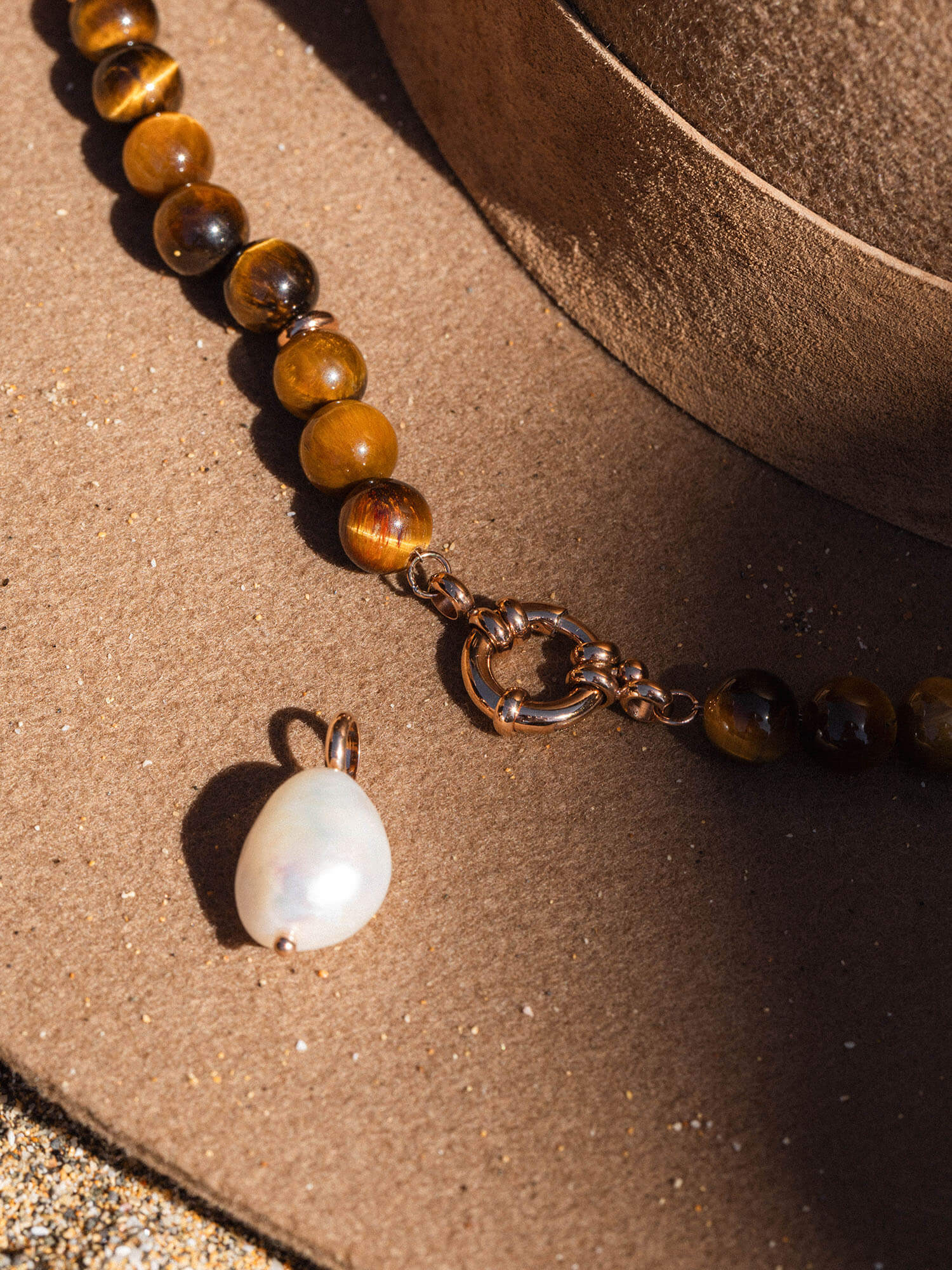 Braune Perlenkette mit Muschel-Anhänger, inspiriert von PURELEI, auf sandigem Untergrund.