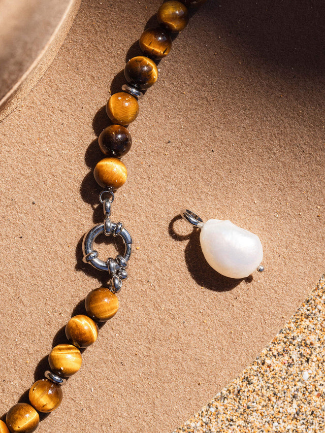 Braune Perlenkette mit silbernem Verschluss und Muschel-Anhänger von PURELEI auf sandigem Untergrund.