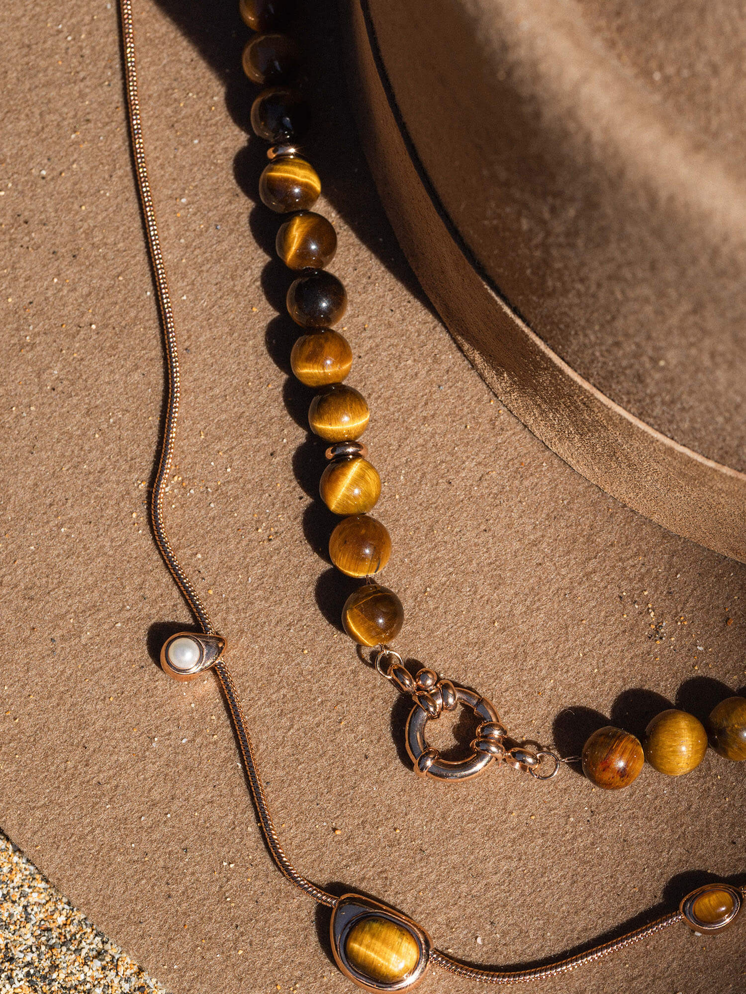 Braune Perlenkette und feine Silberkette auf sandigem Untergrund, inspiriert von PURELEI.