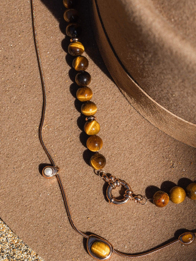 Braune Perlenkette und feine Silberkette auf sandigem Untergrund, inspiriert von PURELEI.