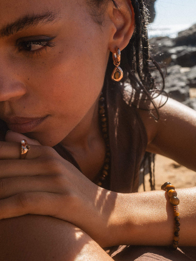 Goldene Ohrringe und Ring von PURELEI, getragen am Strand, strahlen tropische Eleganz aus.