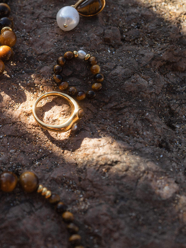 Elastic Anxiety Tiger‘s Eye Pearl Ring mit goldbraunen Tigeraugeperlen und zarter Süßwasserperle auf dunklem Stein.