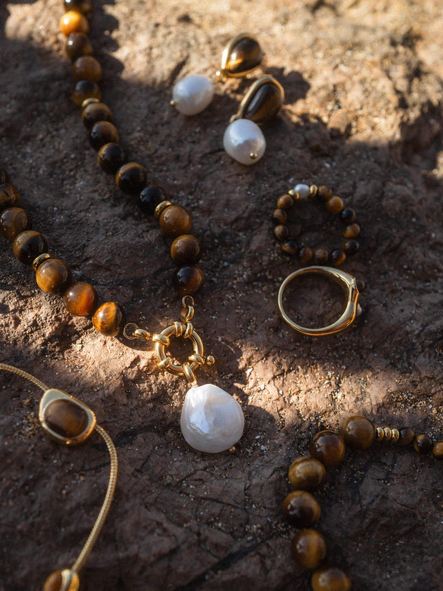 Goldene Schmuckstücke mit Tigerauge und Muschel, elegant auf dunklem Stein arrangiert – PURELEI.
