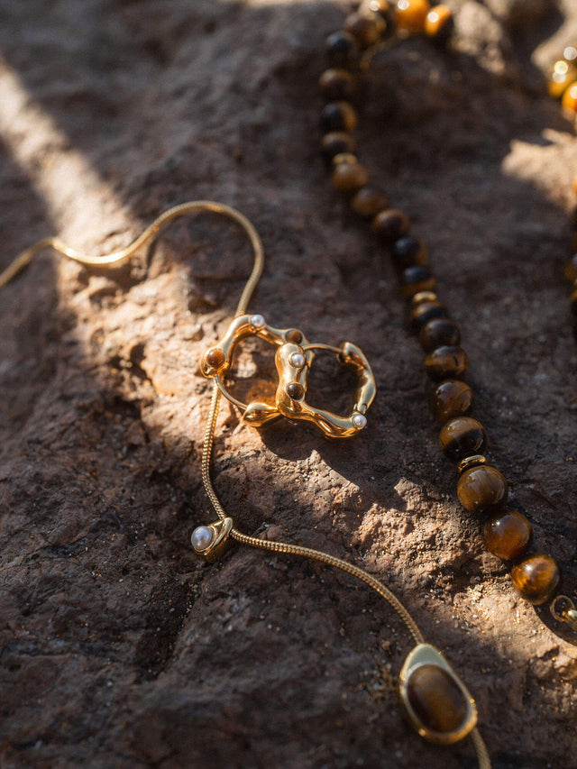 Goldene PURELEI Kette mit organischem Design, umgeben von dunklen Steinen auf rauem Untergrund.