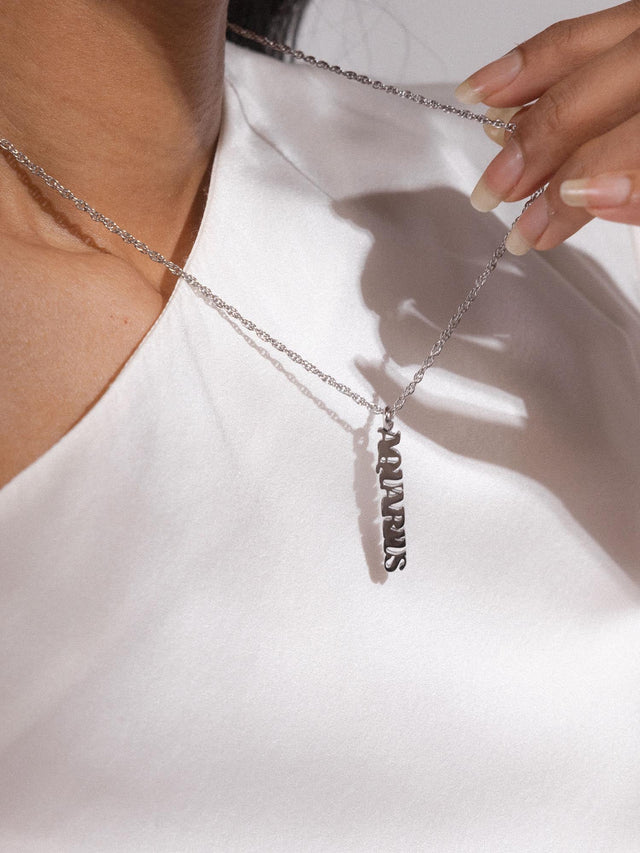 Silberne PURELEI Aquarius Letter Charm Kette mit feinem Schriftzug, elegant und klar im warmen Licht auf weißer Seide.