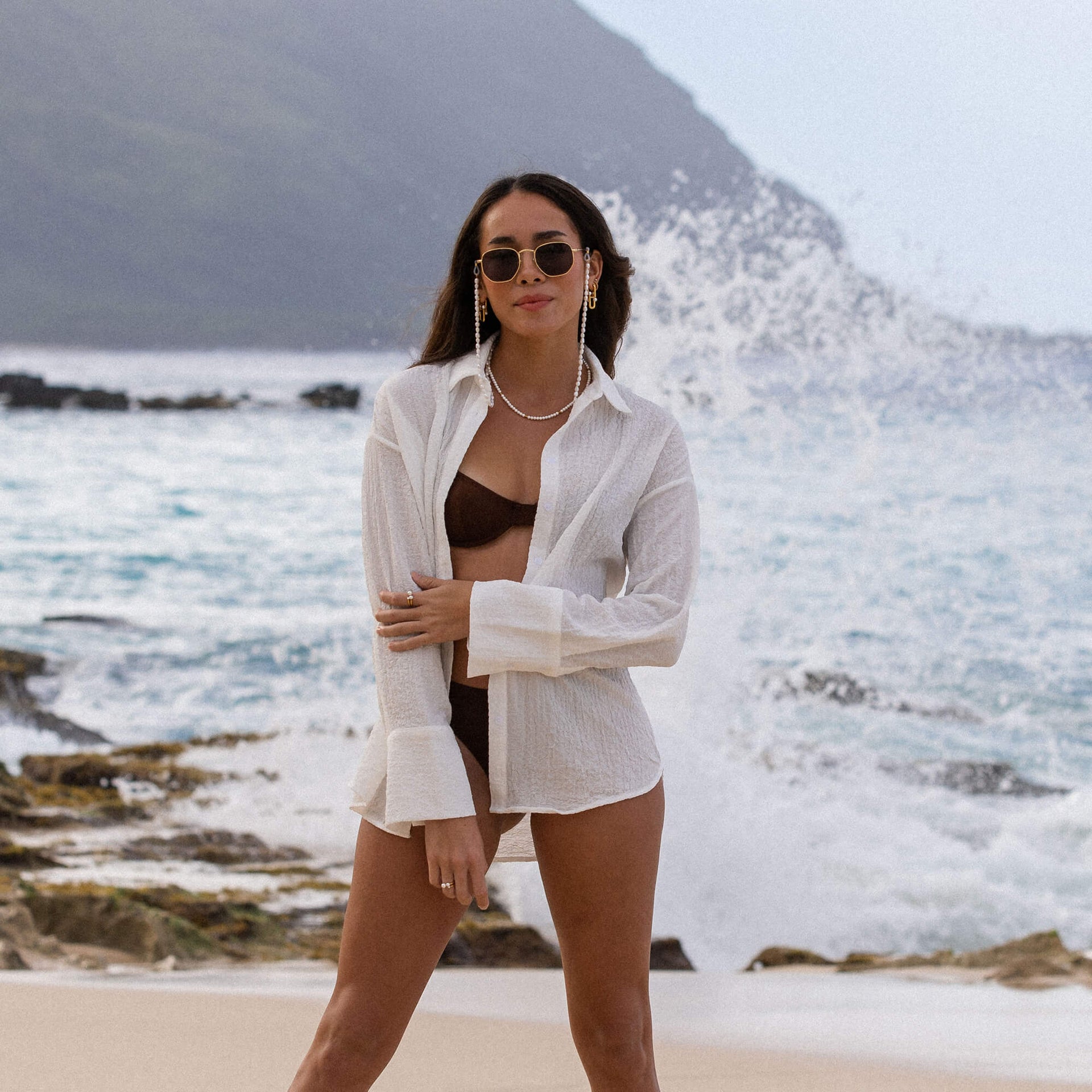 Feminine, weiße Bluse am Strand, kombiniert mit schwarzem Bikini und PURELEI Schmuck.