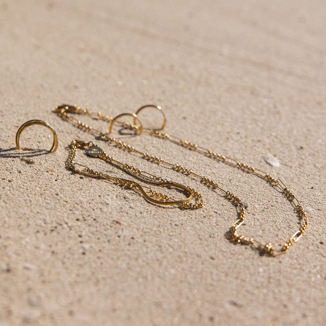 Goldene Kette und Ringe auf Sand, PURELEI inspiriert von tropischer Eleganz.