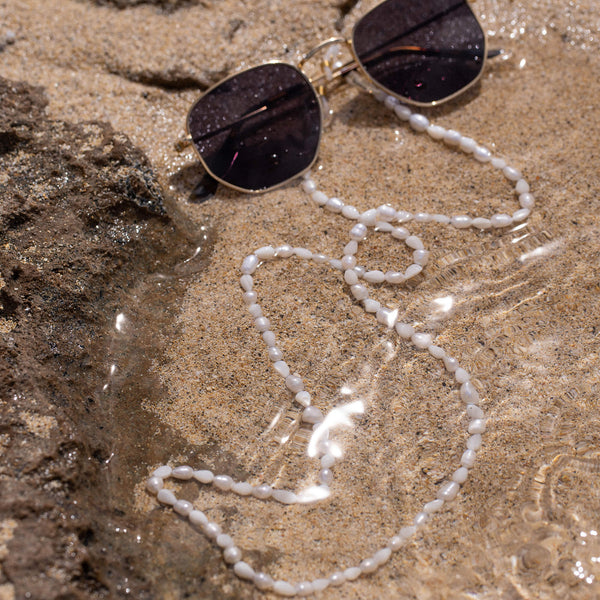 Weiße Muschel-Kette von PURELEI auf Sand, umgeben von Wasser und Sonnenlicht.